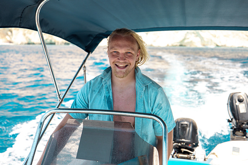 Happy adult caucasian blonde male smiling while driving his cool boat on his vacations in Greece. He has a boat shade above his head. He is standing while holding the steering wheel on the boat. The water is blue.