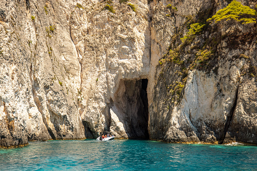 A majestic cliff wall with a big entrance into a sea cave in Greece. A white tourist boat is entering the sea cave on a beautiful summer day. The cliff wall is illuminated by sun. The water is turquoise and crystal clear. There is natural vegetation on the top of the cliffs.