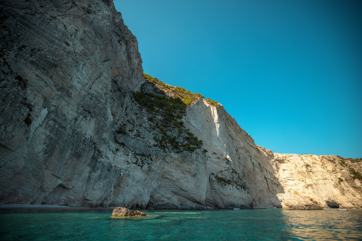 Beautiful high cliffs over the turquoise Ionian sea in Greece. The water is clear and calm. The high cliffs are partly in the shadow. There is a rock  in the middle of the sea protruding through the surface of the water. The weather is nice and sunny.