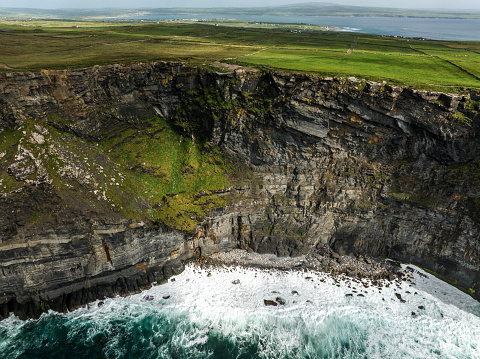 A wild coast on a sunny day with steep cliffs and green grass lawns and a white lighthouse in the far background on a warm sunny summer day