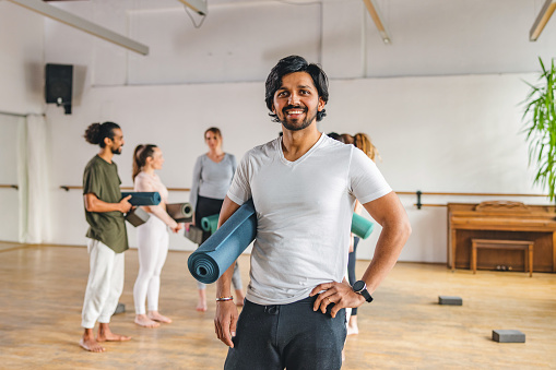A portrait of a Indian yoga teacher posing and smiling at the camera after a yoga session. His clients are having a conversation in the background. They are located in a cosy studio with natural light and wooden floors.