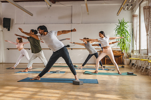 Diverse yoga class participants following the lead and instructions of an Indian male yoga teacher. The yoga class is taking place in a cute vintage studio with wooden floors and big windows. The Space is bright and illuminated by sunrays.