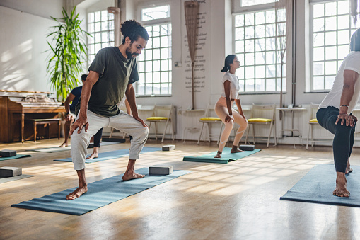 A group of multiracial yoga practitioners standing with their legs wide and knees bent. They are stretching at the start of the yoga class. They are located in a beautifully illuminated yoga studio with big windows.