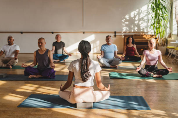 Group Of Multicultural Yoga Participants Seated Meditating Full length front shot with blurred background of multicultural mature yoga participants seated on their mats meditating while following her female instructor indoors during a sunny day. yoga meditation pose stock pictures, royalty-free photos & images