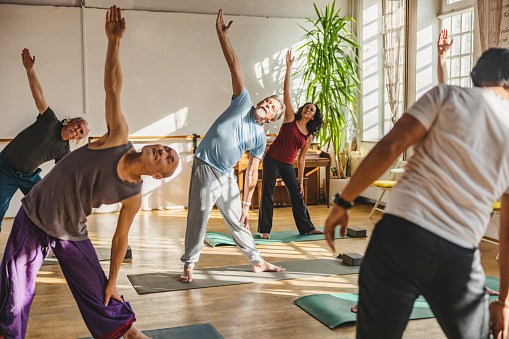 Full length front shot with blurred background of a group of mature Multicultural yoga students practicing a posture while observing and following their male Indian instructor during a sunny day indoors.