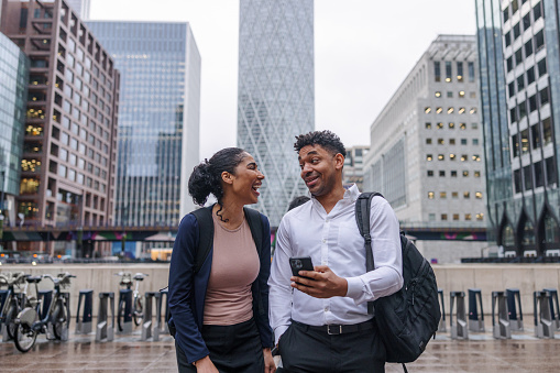 Two dark-skinned coworkers are standing together outside, while one of them is holding a cellphone and looking with surprise at his female colleague next to him.