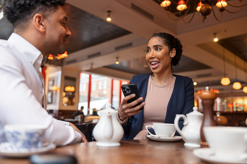 Two dark-skinned colleagues are sitting on the table of a Café and having some coffee and tea together. Over the table there are some ceramic cups and kettles where some tea and coffee is served.