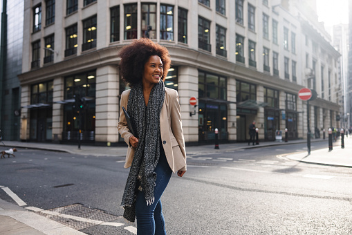 A happy adult black female strolling around the city of London on a sunny winter day. She is wearing business casual clothes. The woman is crossing a street with beautiful architecture and buildings. She is looking away while smiling.