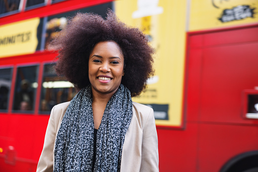 A portrait of an adult black woman looking at the camera and smiling.  She standing on the city street. There is a London double-decker bus in the background. The woman is cheerful and looks happy.