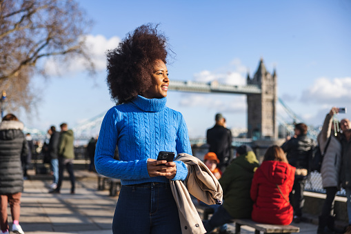 A black adult female tourist happy to be exploring London's landmarks on her own. She is admiring the amazing view of the River Thames near the Tower Bridge. The sun is shining and she is surrounded by many other tourists. She looks happy.