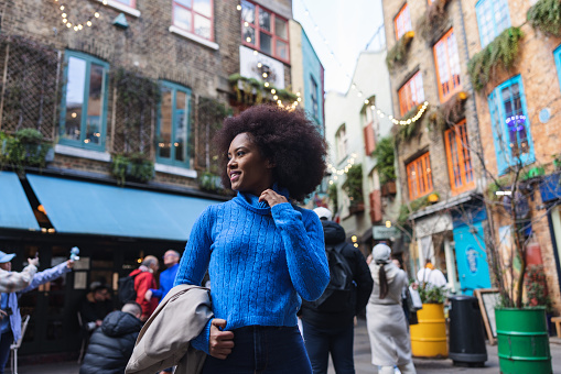 A portrait of an adult black woman exploring the streets of London alone. She is looking away while standing on a colorful street. Her clothes are casual and colorful.