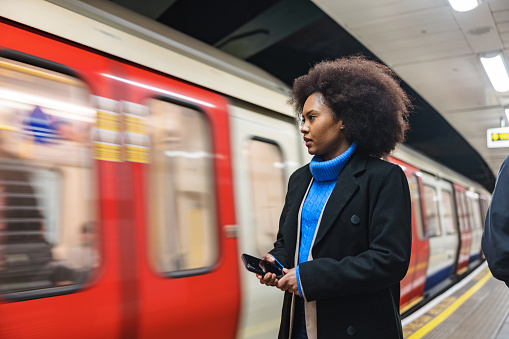 A black adult female waiting on her train at the tube station in London. She is holding a phone in her phone. The woman is on her way home from work. In the background there is a train in motion blur.