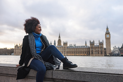 A happy adult black female enjoying the beautiful view of London's landmarks. She is sitting on a railing by the river Thames in front of the Palace of Westminster and Big Ben. The woman is showing her side profile. There is a big smile on her face.