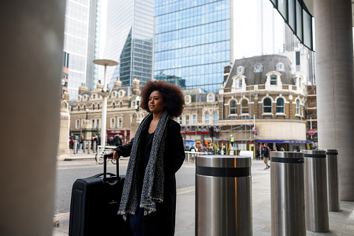 A serious adult black businesswoman with a large suitcase s waiting on a sidewalk for a taxi that will take her to a hotel. She is located in London and is on a business trip. The woman is traveling alone.