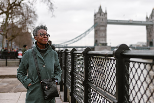 A senior adult black female pensioner enjoying her daily walk by the river Thames in London. She is walking by the railing on the river bank. In the background, the Tower Bridge is partly visible. She looks content and relaxed. The weather is cold and gloomy.