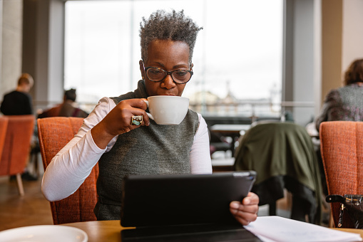 A senior adult black businesswoman drinking coffee at a cafe while working remotely. She is analyzing her business numbers on her digital tablet. The woman is located in a cozy cafe with big windows. The weather is gloomy and typical for London.
