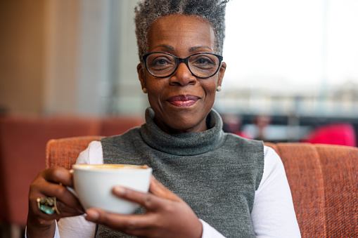 A senior adult black female pensioner smiling at the camera while enjoying her morning coffee at a cosy cafe in London on a gloomy day. She looks content while holding the cup of delicious coffee in her hands.