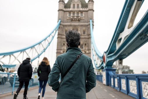 Back view of a senior adult black female walking on a sidewalk and crossing the tower bridge. The woman is wearing a warm emerald green winter coat. The sky is cloudy and the weather is cold.