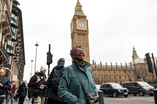 A senior adult black female tourist happy to be visiting London. She is walking around and smiling while admiring the beautiful city. She is located in the City of Westminster. In the background, there is the famous Elizabeth Tower.