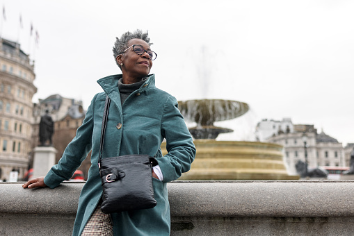 A senior adult black female tourist admiring the architecture in the City of Westminster. She is leaning on a fountain in Trafalgar Square and looking around. The woman has a calm face and looks relaxed. Her clothes are warm since the weather in London is cold and cloudy.