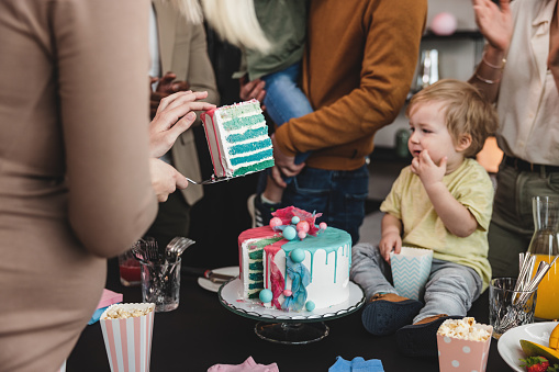 A young Caucasian expectant mother holding a slice of blue cake to reveal the gender of the baby to her family and friends. The people around her are cheering and look happy. Adorable heartfelt moment at the gender reveal celebration.
