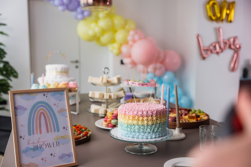 A beautifully decorated table before a baby shower surprise party. The table is full of delicious desserts, sweets and fun props. The room is full of colourful balloons and other decorations. Aesthetic baby shower celebration.