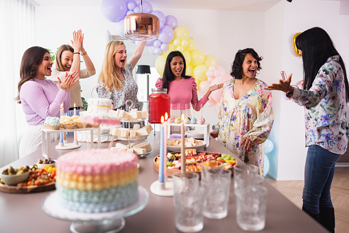 A group of diverse female friends organized a surprise baby shower for their pregnant friend. A beautiful heartwarming gesture. They decorated her modern apartment with balloons and other baby shower decorations. The table is full of delicious sweets and snacks. Everyone is smiling at look happy.