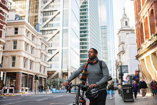 An adult black male admiring the city while walking home from the office. He is pushing his bike while admiring the city on a beautiful sunny day. The man is wearing warm business casual clothes. In the background, there is a street with beautiful buildings that are illuminated by the sunlight .