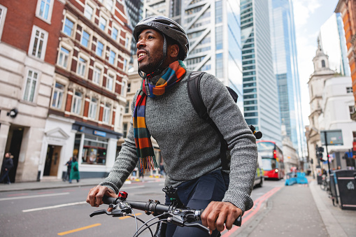 An adult black male using a bike as a transportation home from the office.  He is happy and smiling while biking home. The man is wearing a helmet for safety in the traffic. He is also carrying a backpack.