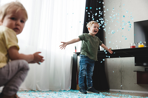 A young caucasian boy playing with blue confetti at a gender reveal party while his younger brother is looking at the camera. Siblings having fun and throwing around confetti.