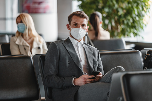 A confident Caucasian businessman wearing a protective face mask, sitting and waiting at the airport lounge. He is looking at the camera.