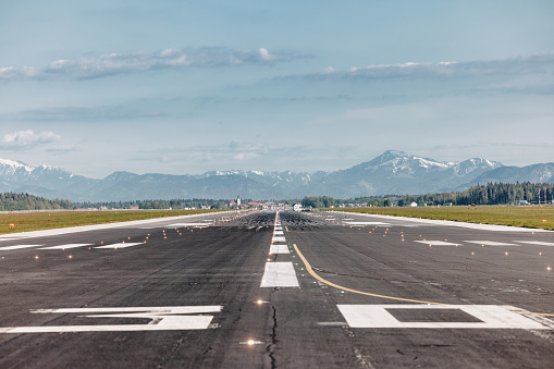 Photo of an empty asphalt airport runway with markings, hills, and mountains in the background.
