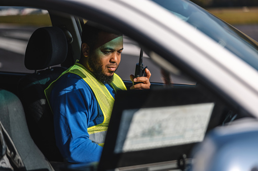 Adult hispanic male who is an airport worker is sitting in work car and listening instructions on walkie talkie. He is very focused. He is wearing blue shirt and yellow safety vest. There is computer set on his passenger seat. Focus is on airport worker.