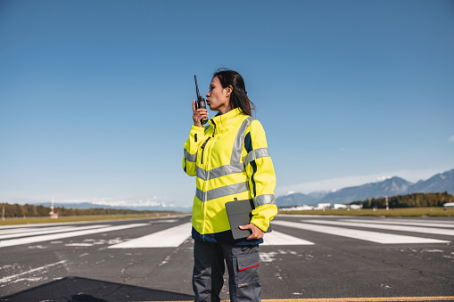 Adult female who is an airport worker is talking on walkie talkie while standing in the middle of an airstrip. She also has electronic tablet device in her other hand. She is looking sideways. She is wearing yellow safety jacket and grey work pants. There are mountains in the background.