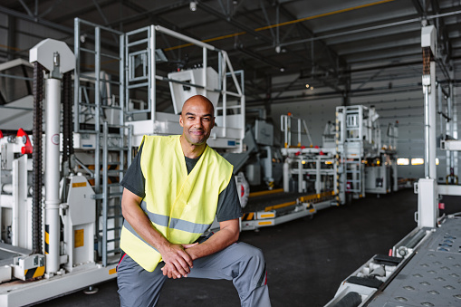 Three-quarter shot with blurred background of a mid-adult Latin-American aeronautic engineer smiling and wearing a green and gray protective uniform as a part of his routine at the warehouse of the airport.