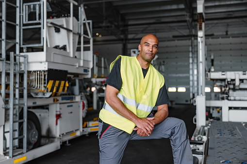 Three-quarter shot with blurred background of a mid-adult Latin-American aeronautic engineer looking serious to the camera and having his arms slightly crossed while wearing a green and gray protective uniform as a part of his routine at the warehouse of the airport.