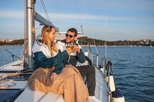 Three-quarter shot with defocused background of a young Caucasian couple sitting on top of the cabin toasting for their anniversary with beer and enjoying the sunset, while the blonde woman has her legs covered with a beige blanket, on a summer day in the Adriatic sea and surrounded by the ropes of the ship.
