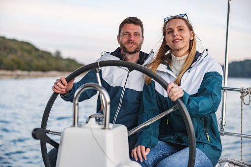 Three-quarter shot with blurred background of a lovely and casually dressed young Caucasian couple holding the helm of the boat while sailing on the Adriatic sea for the weekend as an anniversary celebration.