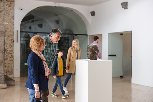 A senior caucasian adult couple visiting a museum on a weekend. They stopped by a bronze statue of a mans head in the museum lobby. They look fascinated and intrigued. Other visitors are walking through the lobby behind the couple.
