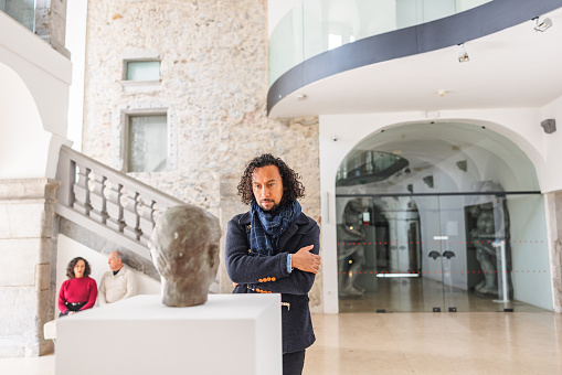 A focused adult black male museum visitor admiring a statue of a man's head . The statue is located on a big white pedestal in the centre of the museum lobby. The man is wearing classy clothes and is standing really close to the statue in order to see better. He is serious as he is looking at the bust. Behind there is a senior couple sitting on the stone bench. The museum is beautiful and bright. It has a stone wall and a stone staircase.