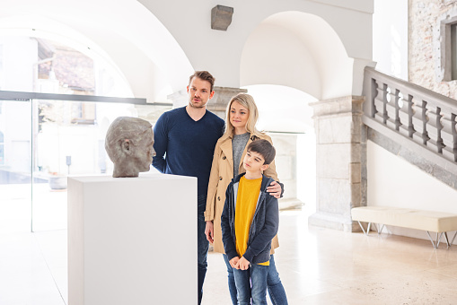 Proud caucasian parent and their young son visiting a museum together on a beautiful morning. They are standing next to a bust on a white pedestal and admiring it. They are having a conversation and look focused. They are standing in a beautiful bright museum with stone details.