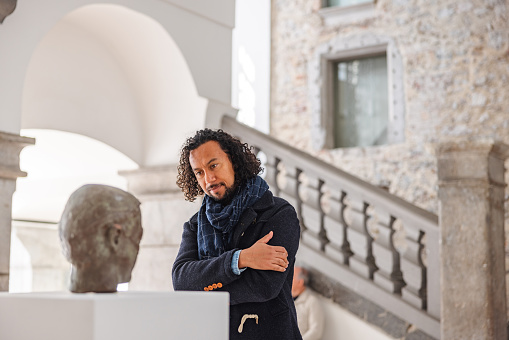 An adult black male history enthusiast standing close to a statue in a history museum and admiring it. He has his arms crossed and is carefully inspecting the statue on the white pedestal. He is located in a beautiful old but renovated museum. He has long curly hair and is wearing a scarf.