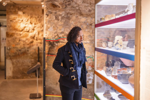 Adult Black Male Museum Visitor Admiring The Old Artefacts In The History Museum A serious adult black male museum visitors admiring the old pottery vessels displayed in the history museum glass display cabinets. He looks interested and intrigued. The man is wearing a scarf and a coat. His hair is long, dark and curly. historical museum stock pictures, royalty-free photos & images