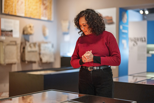 Adult caucasian woman with curly hair standing in a museum and looking at artefacts that are displayed in glass display case. She is focused and serious.