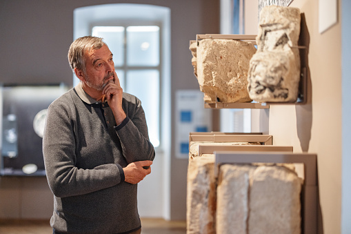 An intelligent senior caucasian male studying the rocks displayed in a history exhibition. He is standing by the display of the stone used for building in the medieval and Roman ages. The man has his hand on the chin while he is thinking. He is serious and focused.