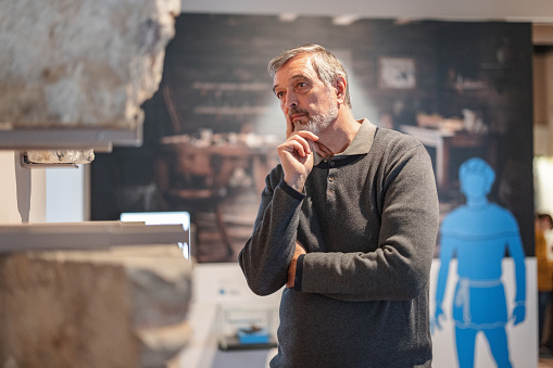 A serious senior caucasian male museum visitor standing in a history exhibition and and looking at a rock that is displayed on the wall. The stones were used to build medieval  buildings. He is standing close in order to see and study it better. he has a thinking but intrigued face. His hand is on his chin as he is thinking.