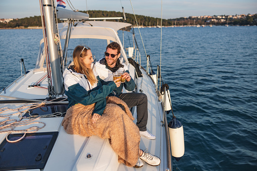 Full length shot with defocused background of a young Caucasian couple sitting on top of the cabin toasting for their anniversary with beer and enjoying the sunset, while the blonde woman has her legs covered with a beige blanket, on a summer day in the Adriatic sea and surrounded by the ropes of the ship.