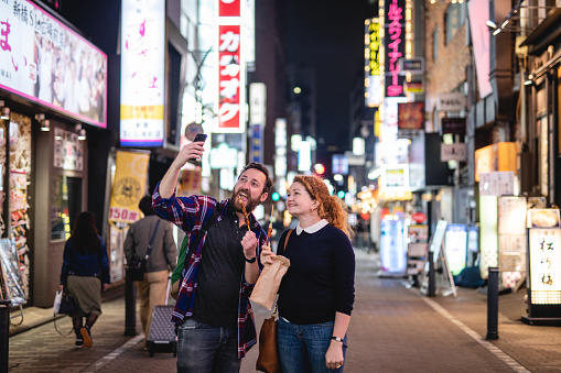 A Caucasian couple smiles and holds food skewers to make a selfie with the cellphone camera. They are standing outside at an illuminated street in Japan at night.