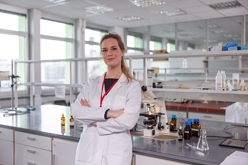 An attractive female young Caucasian scientist wearing a white lab coat and smiling to the camera is standing reclining on the table of a laboratory. She has crossed arms and  behind her there is a microscope and laboratory equipment.