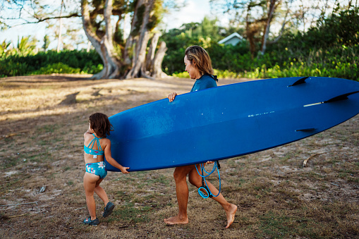 An adorable young girl of Eurasian descent helps her active and healthy grandmother carry a surfboard down the beach to the ocean.